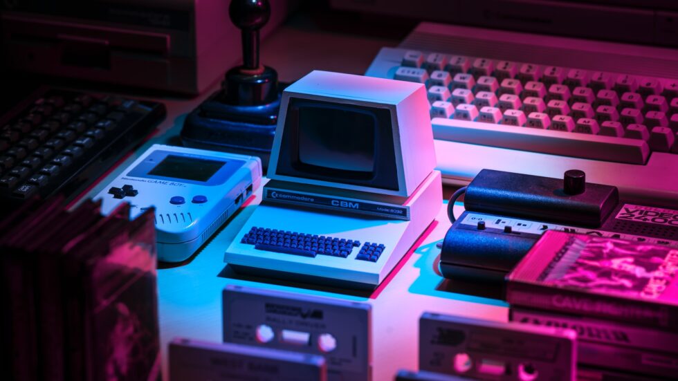 A decorative image that shows a Gameboy, mini Commodore 64, joystick, cassette tapes lit with pink and blue